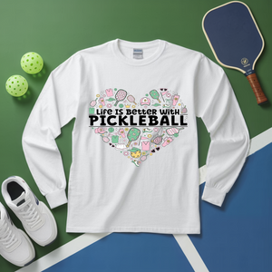 White long-sleeve shirt with pickleball-themed design on a green and blue surface with pickleballs and a paddle.