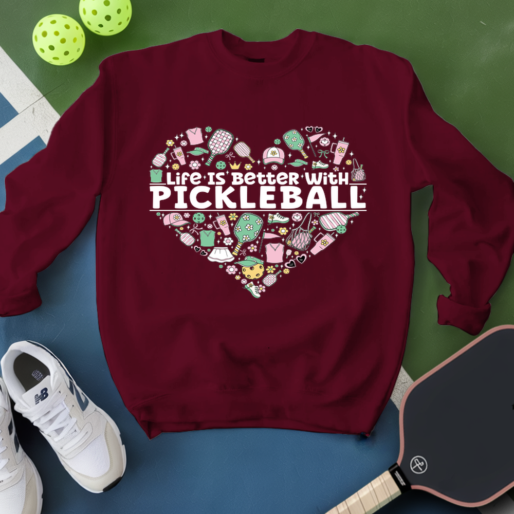 Maroon sweatshirt with pickleball design and text, surrounded by pickleballs and a paddle on a green and blue background.