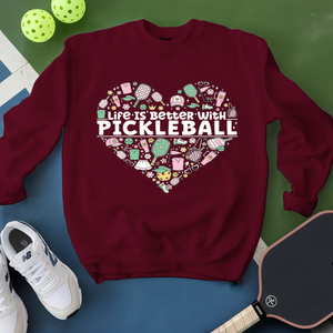 Maroon sweatshirt with pickleball design and text, surrounded by pickleballs and a paddle on a green and blue background.