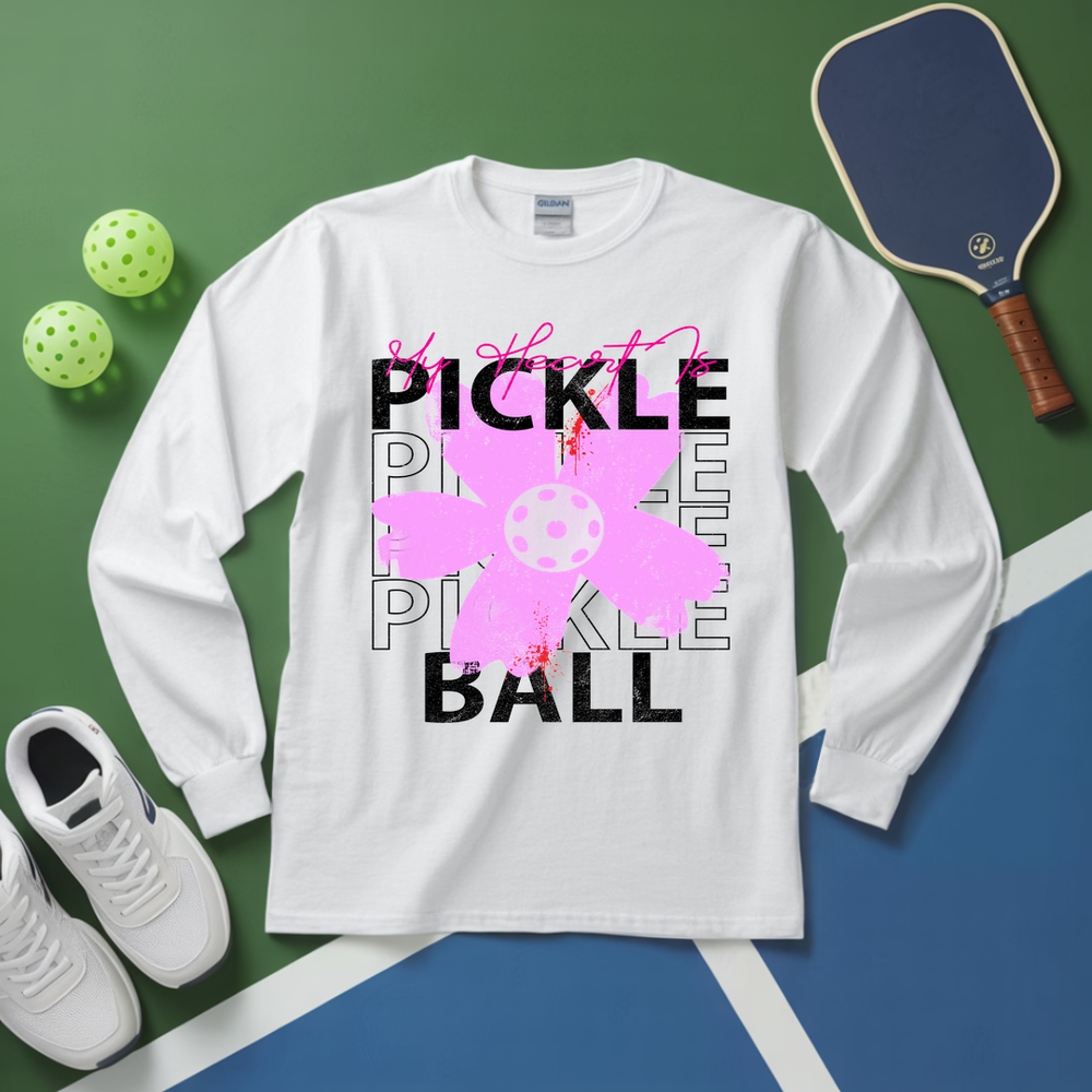 White long-sleeve shirt with pickleball-themed design on a green and blue surface with pickleballs and a paddle.