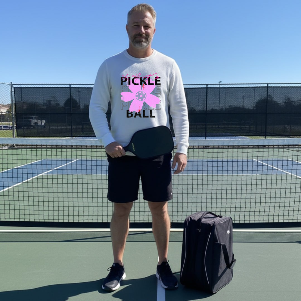 Man on a pickleball court holding a paddle and wearing a sweatshirt with a pickleball ball graphic.