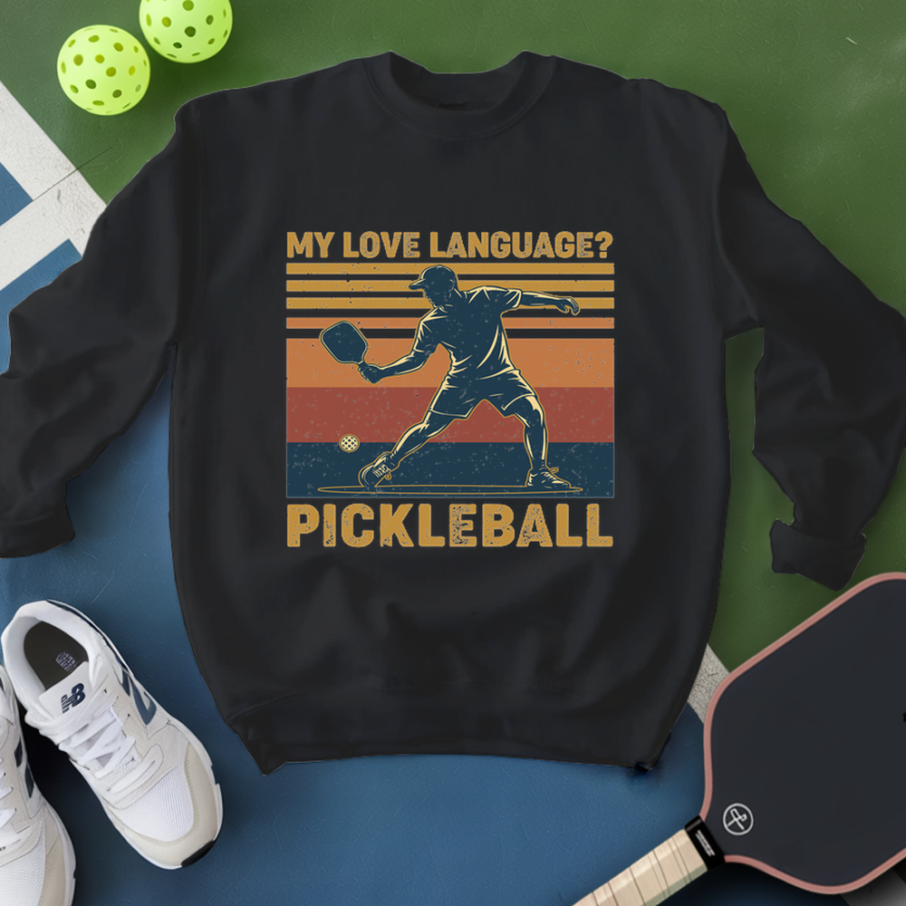 Black sweatshirt with pickleball design and text, surrounded by pickleballs and a paddle on a green and blue background.