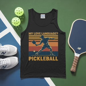 Black tank top with pickleball graphic and text, surrounded by pickleballs, shoes, and paddle on a green and blue surface.