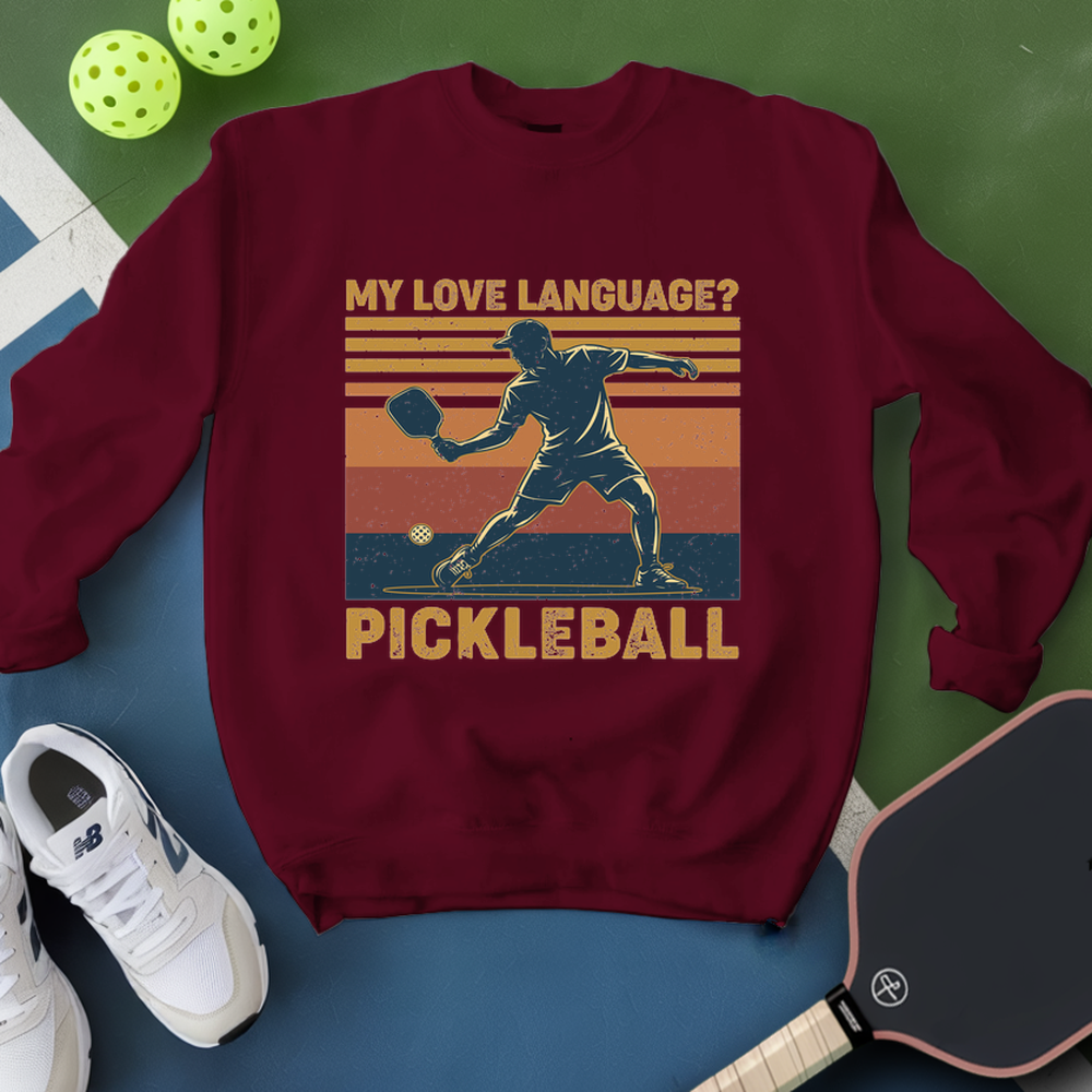 Maroon sweatshirt with pickleball design and text, surrounded by pickleballs and a paddle on a green and blue background.