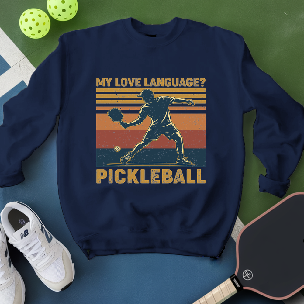 Navy blue sweatshirt with pickleball design and text, surrounded by pickleballs and a paddle on a green and blue background.