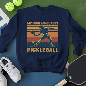 Navy blue sweatshirt with pickleball design and text, surrounded by pickleballs and a paddle on a green and blue background.
