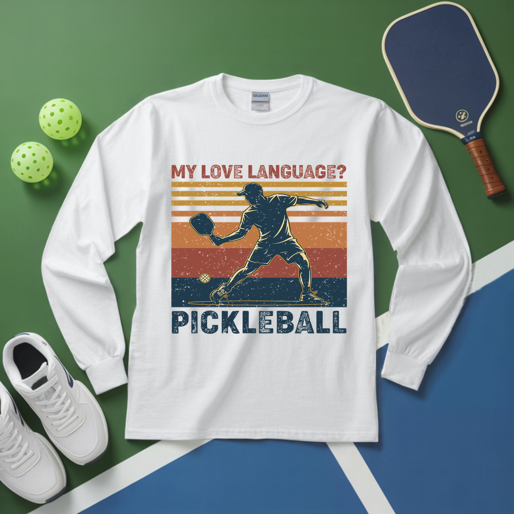 White long-sleeve shirt with pickleball graphic and text on a green and blue surface with pickleball equipment.