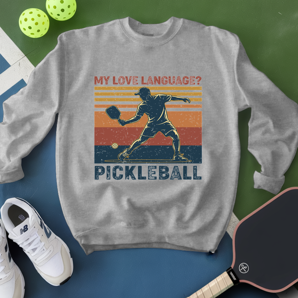 Gray sweatshirt with pickleball design and text, surrounded by pickleballs and a paddle on a green and blue background.