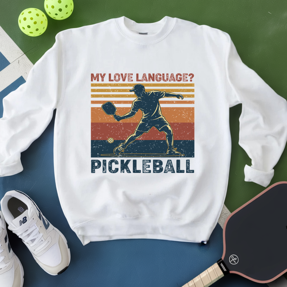 White sweatshirt with pickleball design and text, placed on a green and blue surface with pickleballs and a paddle.