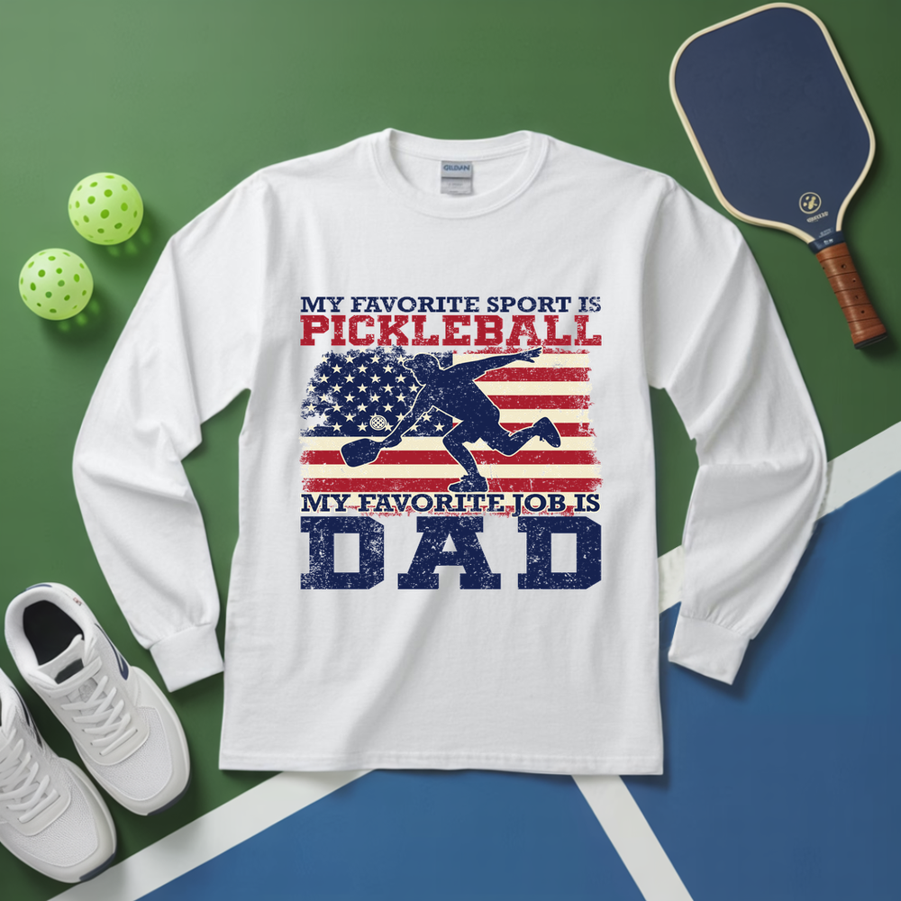 White long-sleeve shirt with pickleball-themed design on a green and blue surface with pickleball equipment.