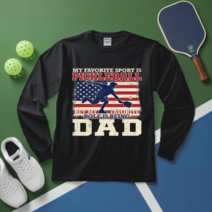 Black long-sleeve shirt with pickleball-themed design on a green and blue surface with pickleball equipment.