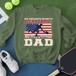 Green sweatshirt with pickleball design and text, surrounded by pickleballs and a paddle on a green and blue background.