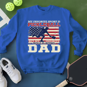 Blue sweatshirt with pickleball design and text on a green surface with pickleballs and a paddle.