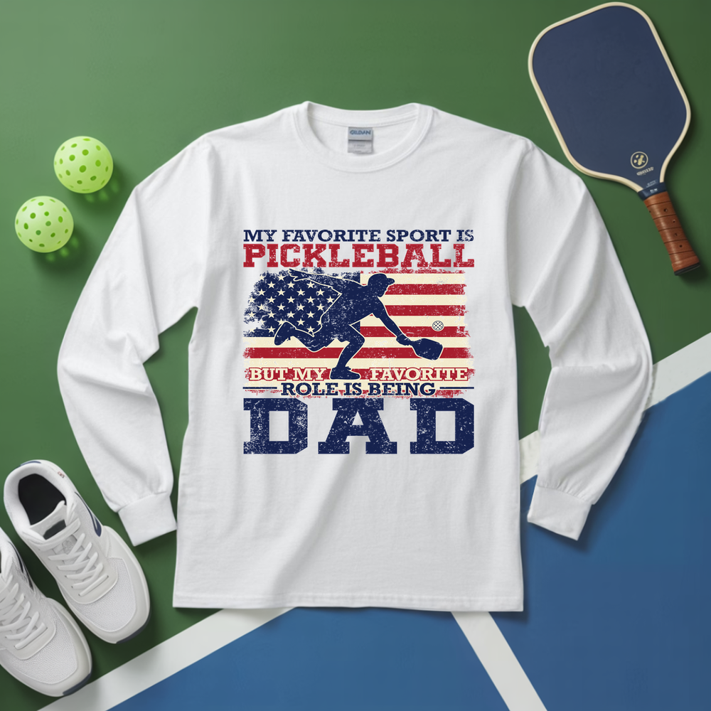 White long-sleeve shirt with pickleball-themed design on a green and blue surface with pickleball equipment.