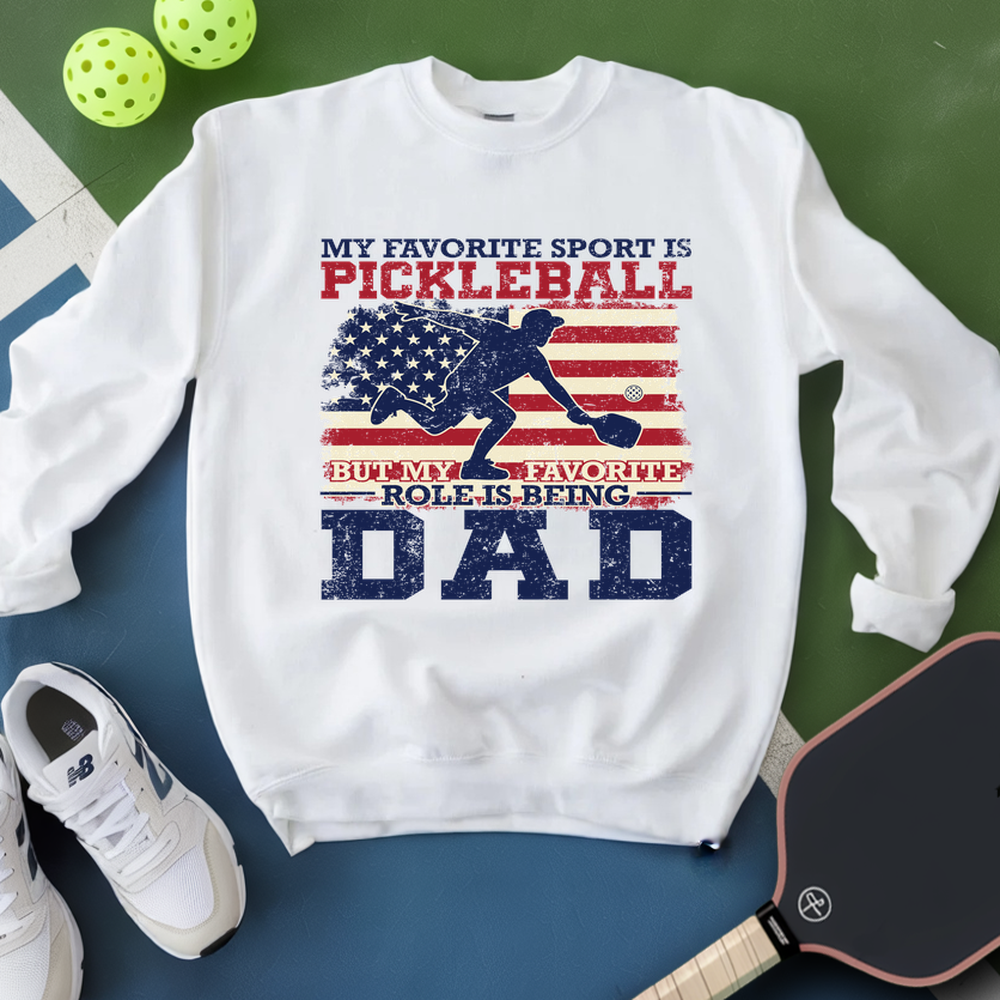 White sweatshirt with pickleball-themed design on a green and blue surface with sports equipment.