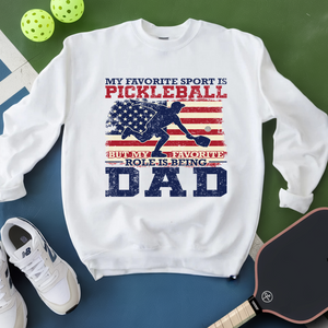 White sweatshirt with pickleball-themed design on a green and blue surface with sports equipment.