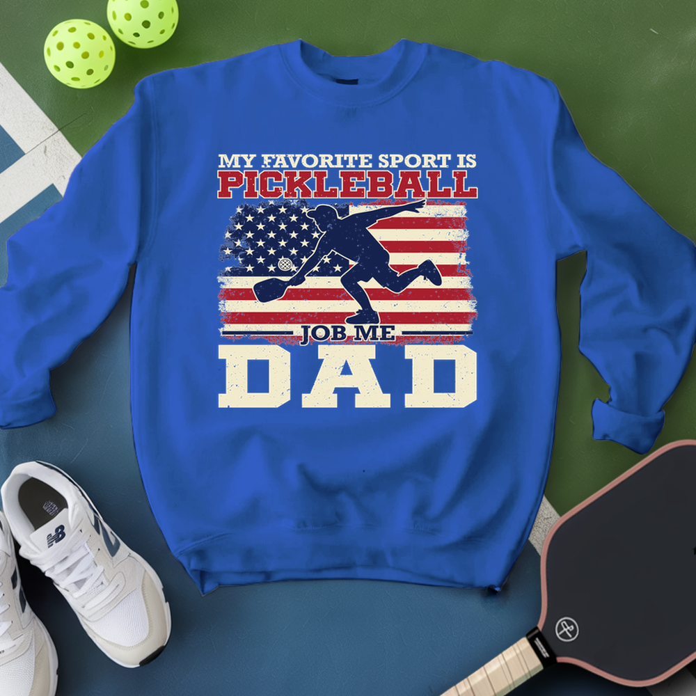 Blue sweatshirt with pickleball-themed design on a green surface with pickleballs and a paddle.