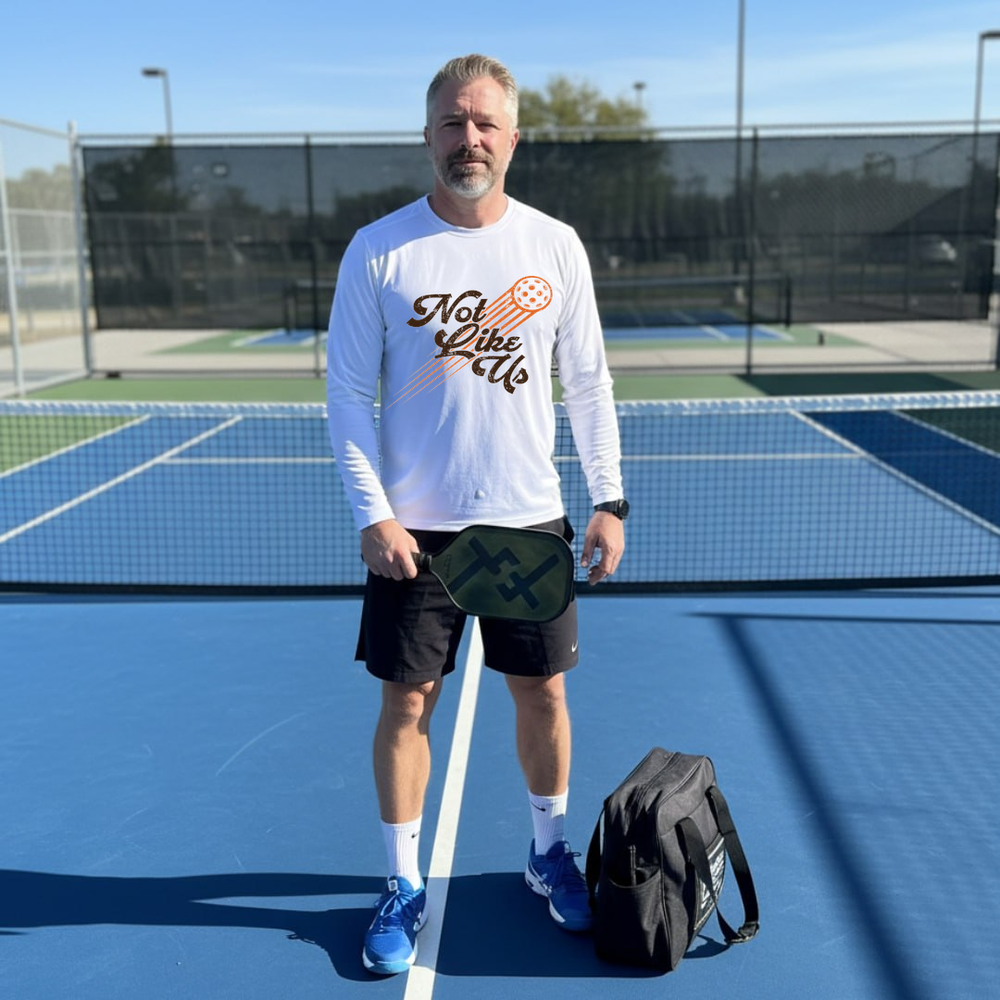 Green long-sleeve shirt with 'Not Like Us' design on a green and blue surface with pickleball equipment.