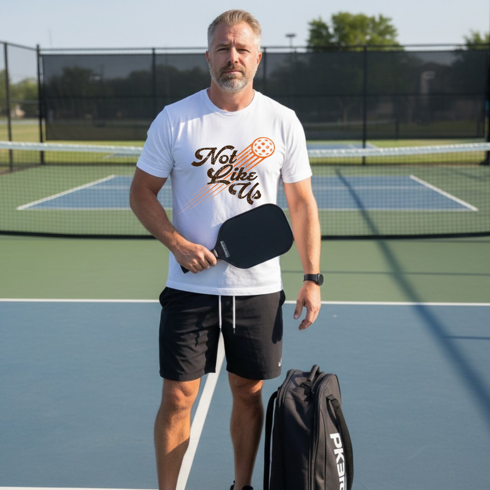 Man standing on a pickleball court holding a paddle and wearing a white t-shirt with text and graphics.