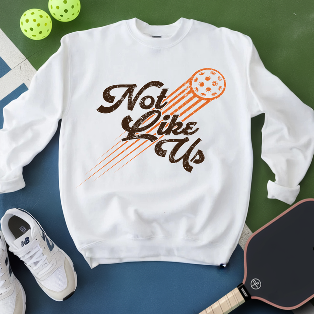 White sweatshirt with 'Not Like Us' graphic, white sneakers, and pickleball paddle on a green and blue background