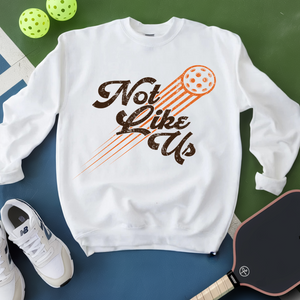 White sweatshirt with 'Not Like Us' graphic, white sneakers, and pickleball paddle on a green and blue background