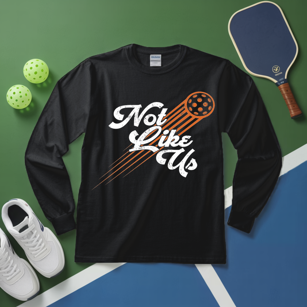 Black long-sleeve shirt with 'Not Like Us' graphic on a sports court background with pickleball equipment.