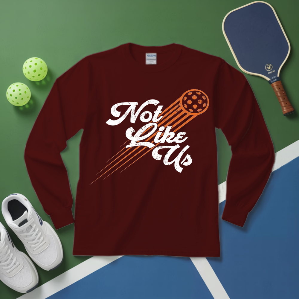 Maroon long-sleeve shirt with 'Not Like Us' graphic on a green and blue surface with pickleball equipment.