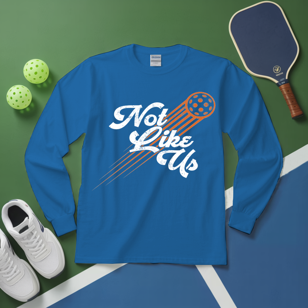 Blue long-sleeve shirt with 'Not Like Us' graphic on a green and blue surface with pickleball equipment.