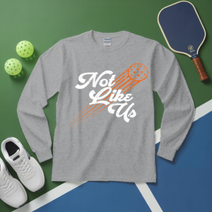 Gray long-sleeve shirt with 'Not Like Us' design on a green and blue surface with pickleball equipment.