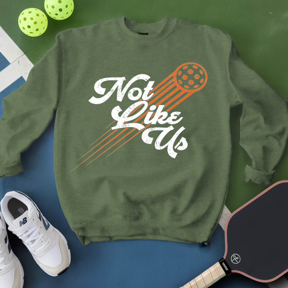 Green sweatshirt with 'Not Like Us' text and pickleball paddle graphic, surrounded by pickleballs and a paddle on a green and blue background.