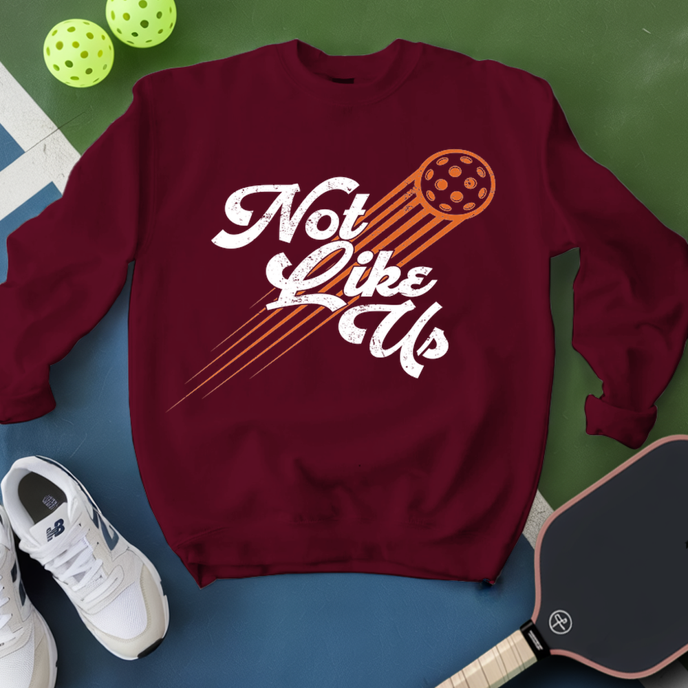 Maroon sweatshirt with 'Not Like Us' graphic on a green and blue background with pickleball equipment.