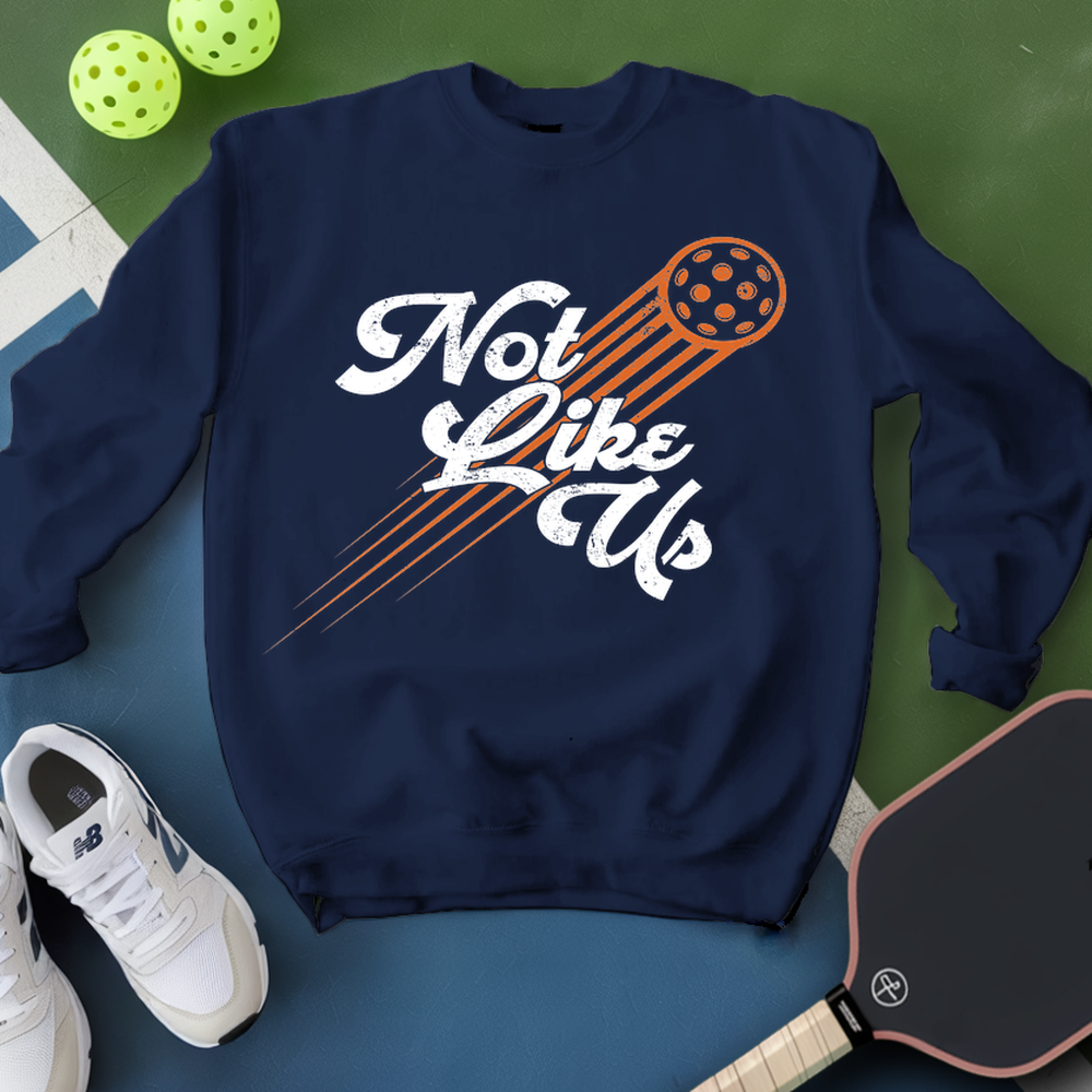 Navy blue sweatshirt with 'Not Like Us' text and pickleball paddle graphic, surrounded by pickleballs and a paddle on a green and blue background.