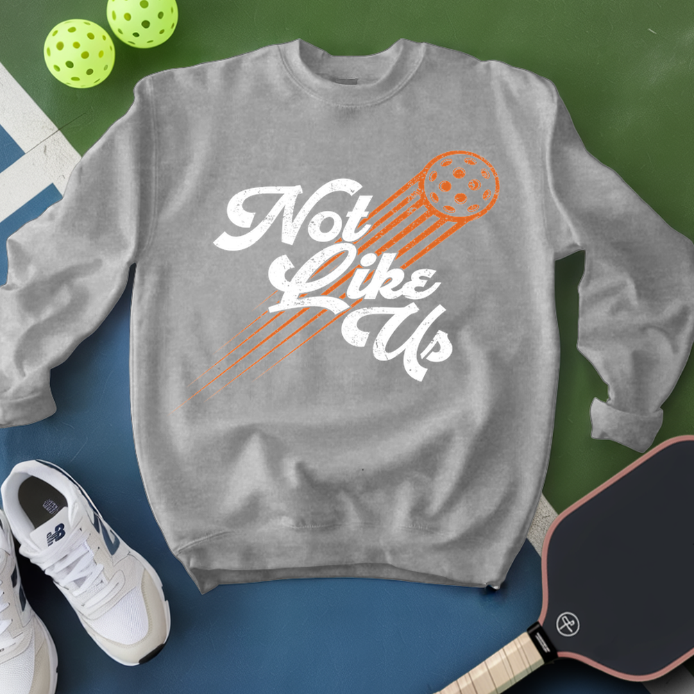 Gray sweatshirt with 'Not Like Us' text and pickleball paddle graphic, surrounded by pickleballs and a paddle on a green and blue background.