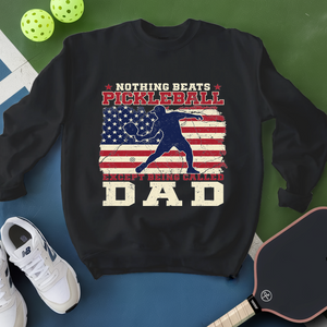 Black sweatshirt with pickleball design and text, surrounded by pickleballs and a paddle on a green and blue background.