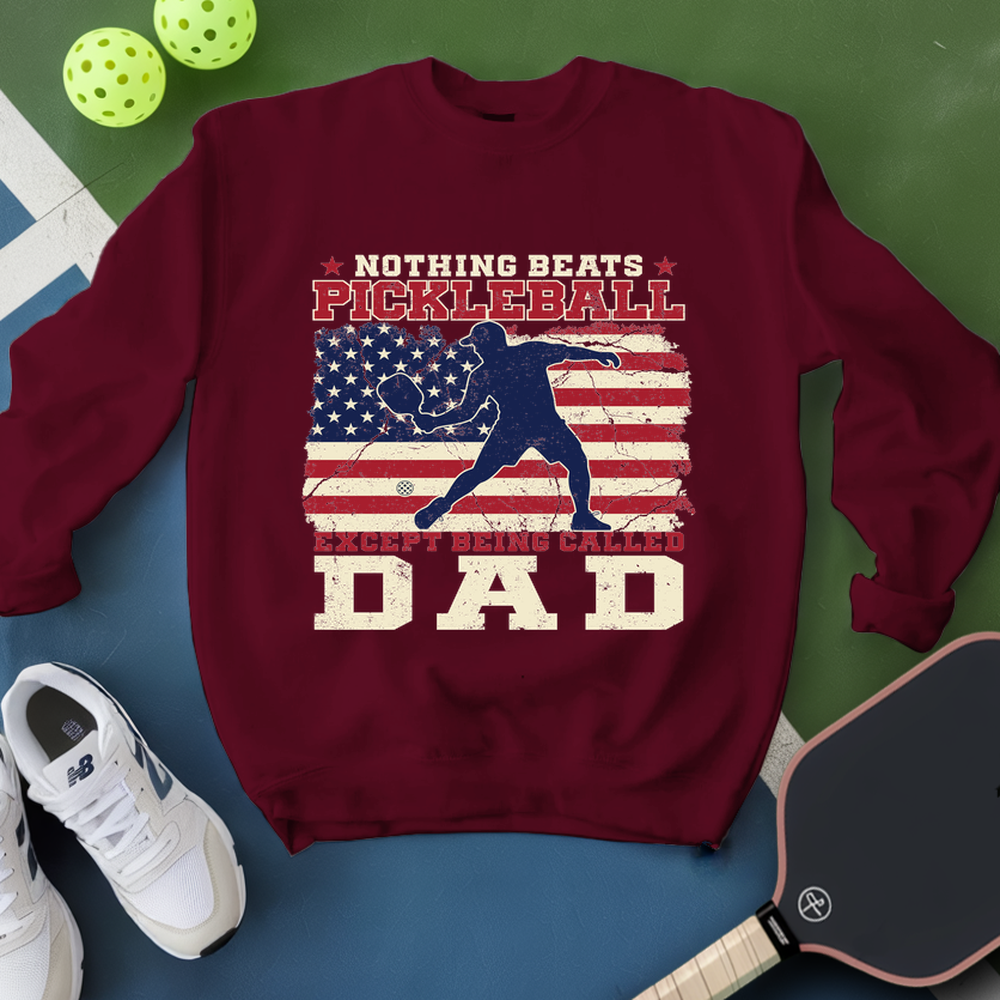 Maroon sweatshirt with pickleball design and text, surrounded by pickleballs and a paddle on a green and blue background.