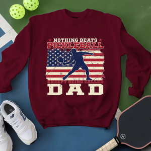 Maroon sweatshirt with pickleball design and text, surrounded by pickleballs and a paddle on a green and blue background.