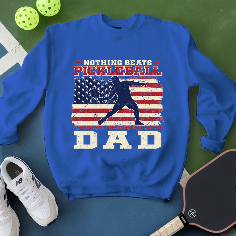 Blue sweatshirt with pickleball design and text on a green surface with pickleballs and a paddle.