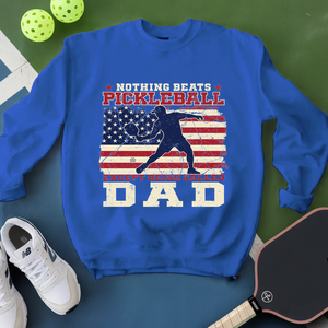 Blue sweatshirt with pickleball design and text on a green surface with pickleballs and a paddle.