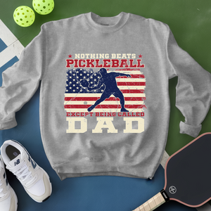 Gray sweatshirt with pickleball design and text, surrounded by pickleballs and a paddle on a green and blue background.