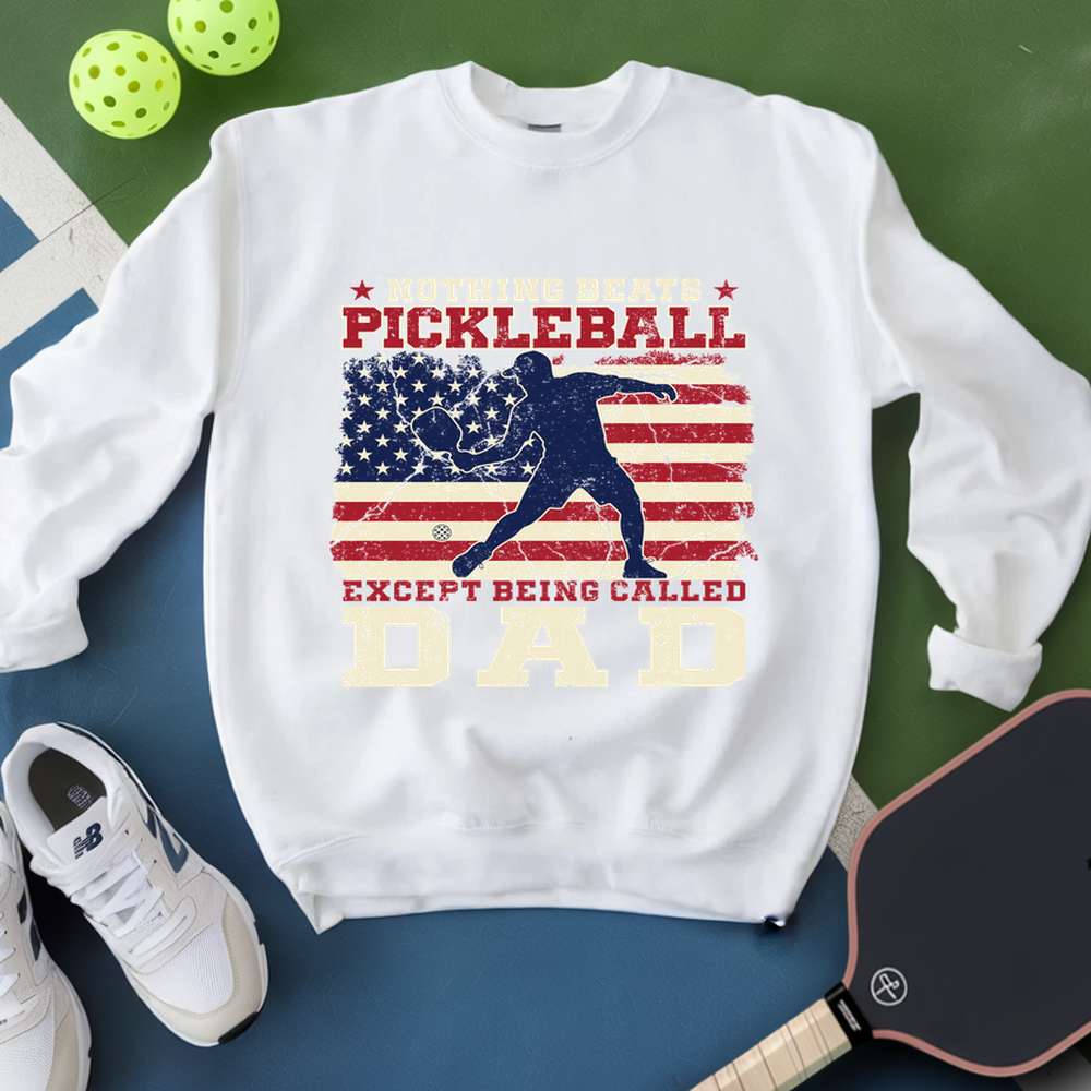 White sweatshirt with pickleball design and text, placed on a green and blue surface with pickleballs and a paddle.