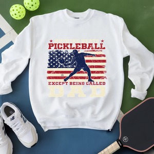 White sweatshirt with pickleball design and text, placed on a green and blue surface with pickleballs and a paddle.