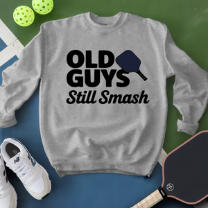 Gray sweatshirt with 'Old Guys Still Smash' text, surrounded by pickleball equipment on a green and blue background.