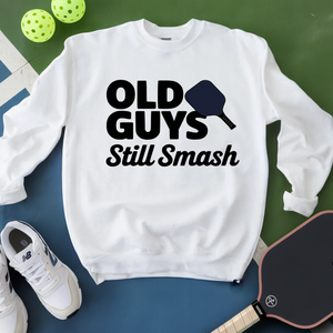 White sweatshirt with 'Old Guys Still Smash' text, pickleball paddle, and balls on a green and blue background