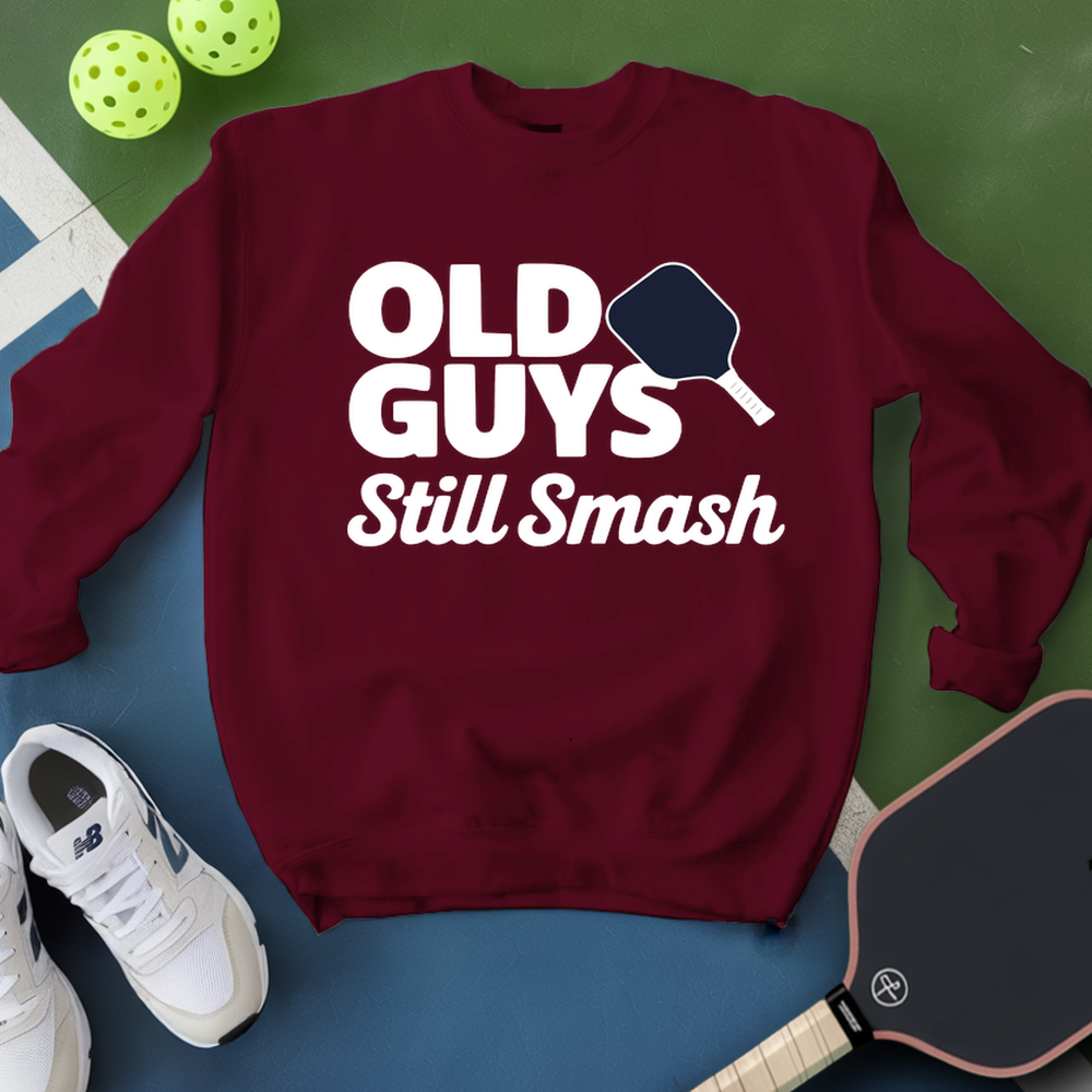 Maroon sweatshirt with 'Old Guys Still Smash' text, pickleball paddles, and balls on a green and blue background