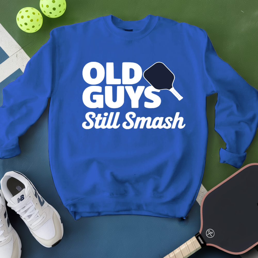 Blue sweatshirt with 'Old Guys Still Smash' text, pickleball paddles, and balls on a green surface