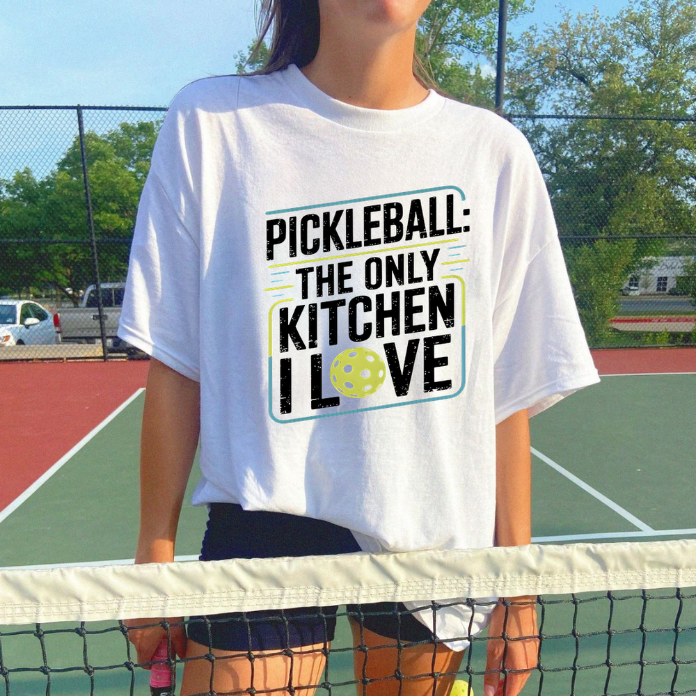 Person wearing a white t-shirt with pickleball-themed text on a tennis court