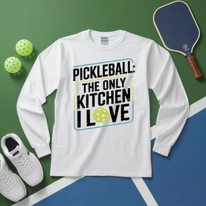 White long-sleeve shirt with pickleball-themed text on a green and blue surface with pickleballs and a paddle.