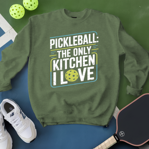 Green sweatshirt with pickleball-themed text, surrounded by pickleballs and a paddle on a green and blue background.