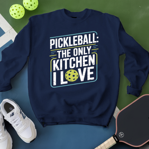 Navy blue sweatshirt with pickleball-themed text, surrounded by pickleballs and a paddle on a green and blue background.