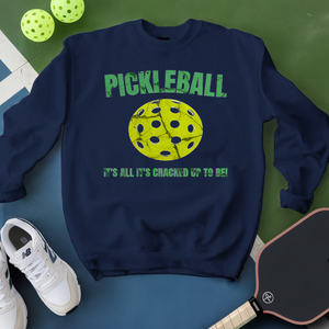 Navy sweatshirt with pickleball graphic and text, surrounded by pickleballs and a paddle on a green and blue background.